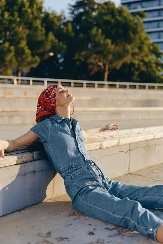 Lifestyle woman in modern boho-western denim street style relaxing outdoors, wearing a red bandana with a warm film color tone, enjoying sunny urban environment.