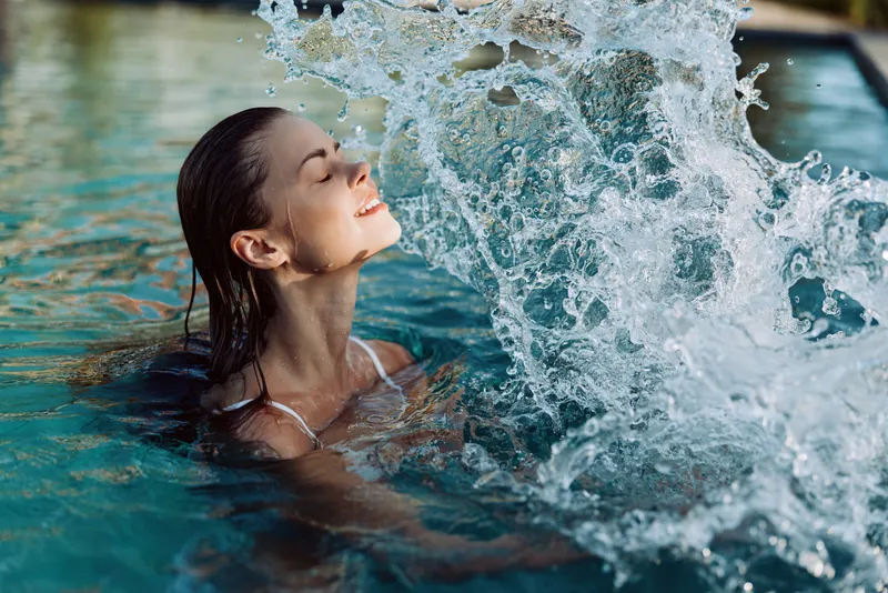 A woman floats in a pool, water spray rising as she tilts her head back, a vivid splash framing her serene face. This scene conveys relaxation, leisure, and wellness in a sunlit outdoor moment.
