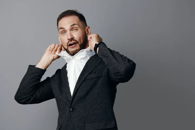 A business professional pulls at his collar against a plain colored backdrop, conveying tension, nerves, and readiness in a moment of decision, ideal for corporate, leadership, or financial industry