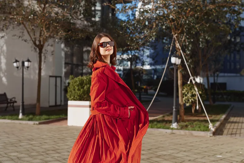 Cheerful woman outdoors in a bright red coat, wearing sunglasses, smiling and relaxed, sunlit street scene with trees and greenery, showcasing a vibrant fashion moment