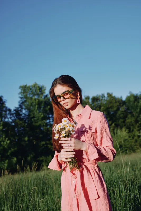 Young woman in a pink dress holding wildflowers, expressing serenity in a lush green field under a clear blue sky