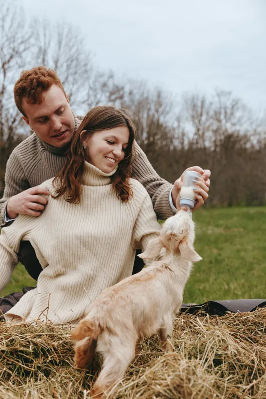 Couple feeds a kid goat with a bottle outdoors, joyful atmosphere, woman smiling, man guiding, cozy sweater, hay bale, grassy rural field, candid lifestyle moment with animals