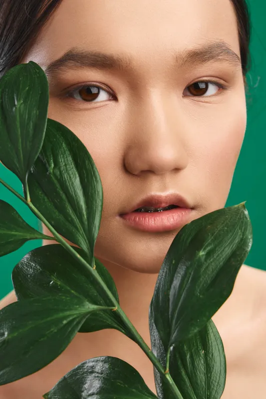 Asian woman poses with glossy green leaves partially framing her face in a minimalist studio setting. The calm expression and soft lighting convey natural beauty and serenity.