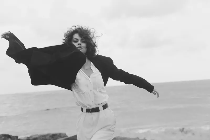 Woman in white shirt and blazer spreads arms on a windy beach, clothing flowing, black and white photography, motion and freedom, fashion mood and dynamic texture that captures style and atmosphere