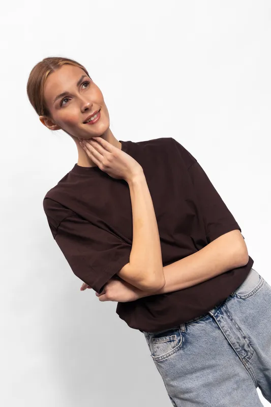 Young woman with light skin and brown hair wearing a dark brown shirt and blue jeans poses thoughtfully against white background. Concept of casual fashion and natural beauty portrait.