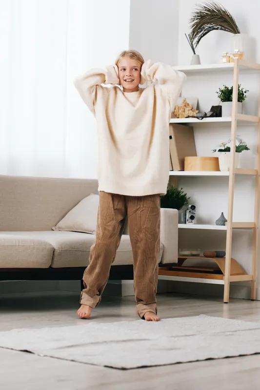 A cheerful child wearing an oversized cream sweater and plush pants stands barefoot in a bright living room, smiling with hands resting behind the head, creating a cozy, inviting scene.