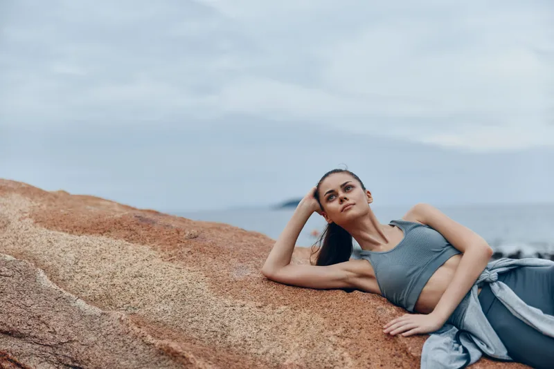 Young woman in activewear lounging on a rocky beach, showcasing a serene expression and relaxed posture, perfect for wellness and fitness themes