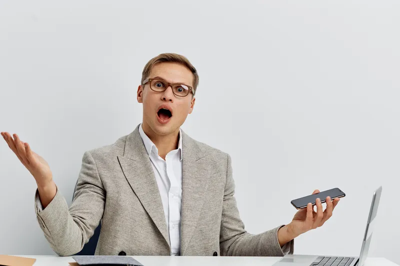 Concerned man wearing business casual outfit with glasses gestures with surprised expression, holding smartphone in hand, sitting at desk with laptop and documents, isolated on plain neutral backgroun