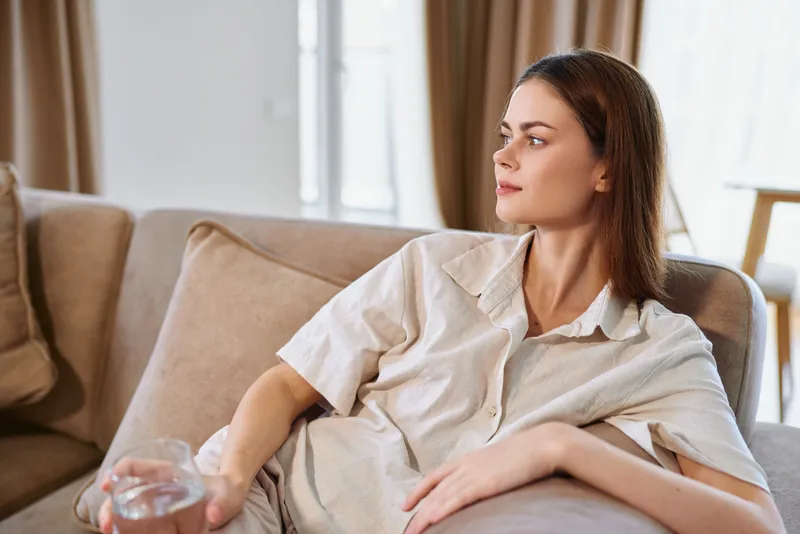Woman on sofa at home in casual clothes, portrait of relaxation and comfort in a livingroom, sitting with a drink in hand, thoughtful gaze toward window, cozy cushions and soft morning light