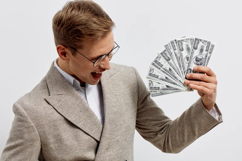 Confident young man with glasses wearing beige blazer holds fan of hundred dollar bills joyful expressively isolated on plain white background studio portrait. Finance success wealth concept.