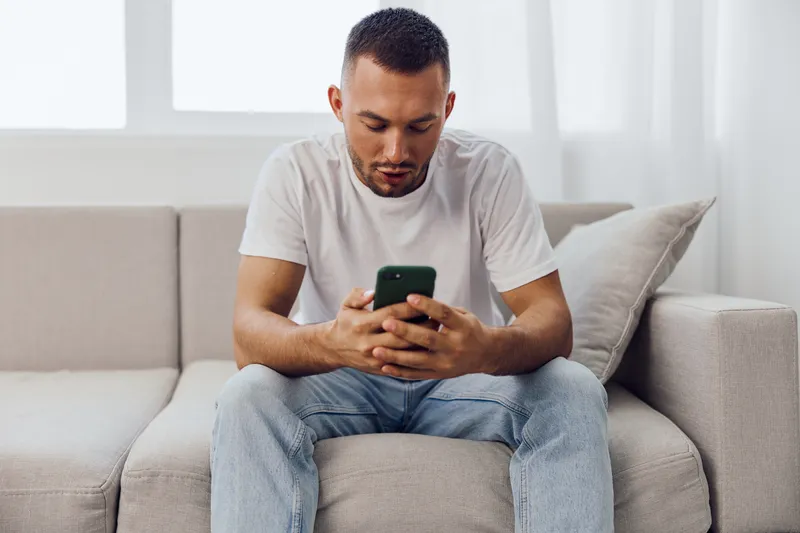 Concentrating man using smartphone while sitting on a comfortable sofa, dressed in casual attire. A moment of modern lifestyle and technology in a bright room.