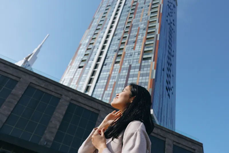 Woman gazing up at a towering glass skyscraper in a modern city, urban architecture and clear sky, conveying ambition, confidence and momentum from a street level perspective