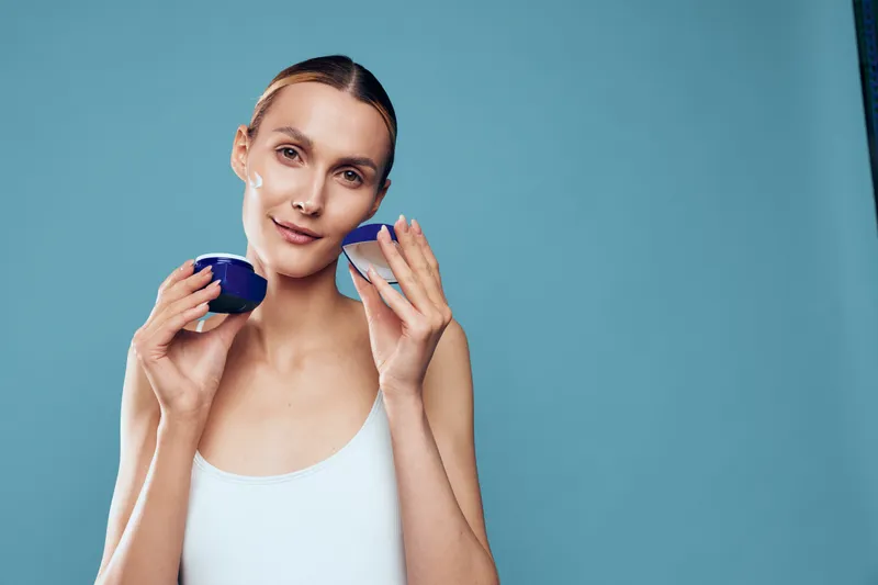 Young woman with clear skin holds open jar of face cream applies moisturizer on cheek isolated on blue background in skincare beauty concept.