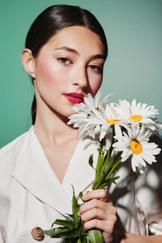Portrait of a young woman with flawless skin and vibrant red lips holding a bouquet of fresh daisies against a soft green background. Concept of beauty, fashion, and elegance.