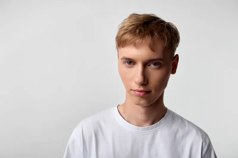 Man with short light hair in a white tshirt stands in a clean studio with neutral background, calm expression, minimal aesthetic for fashion and lifestyle shoots
