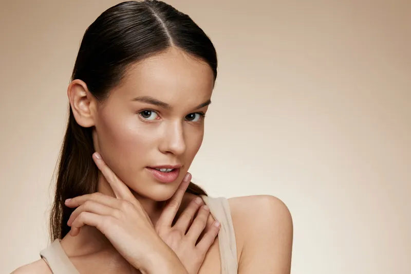 Young woman with a natural look, wearing neutral toned clothing, showcasing confidence and beauty against a soft beige background, ideal for beauty and skincare concepts