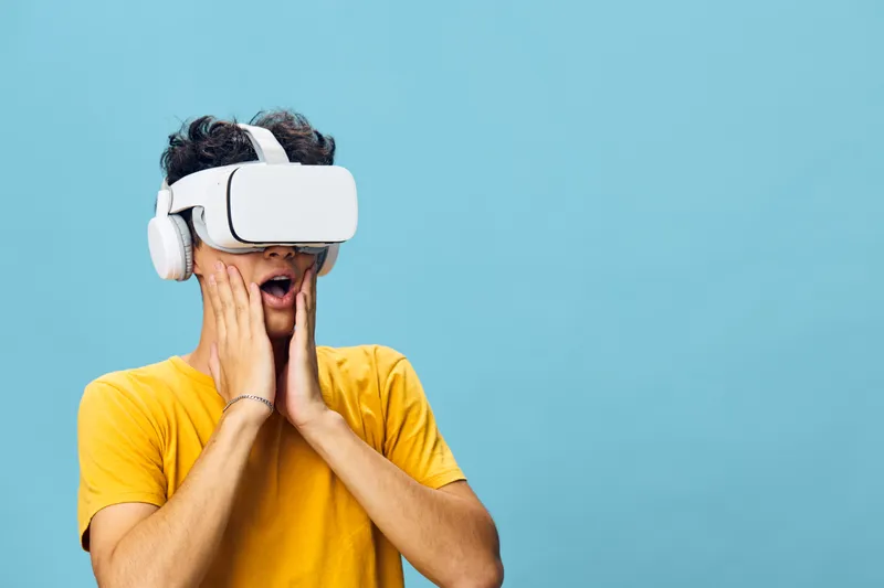 Молодой человек в VR-шлеме, явно удивленный, в изумлении прижимает руки к лицу на фоне мягкого голубого фона