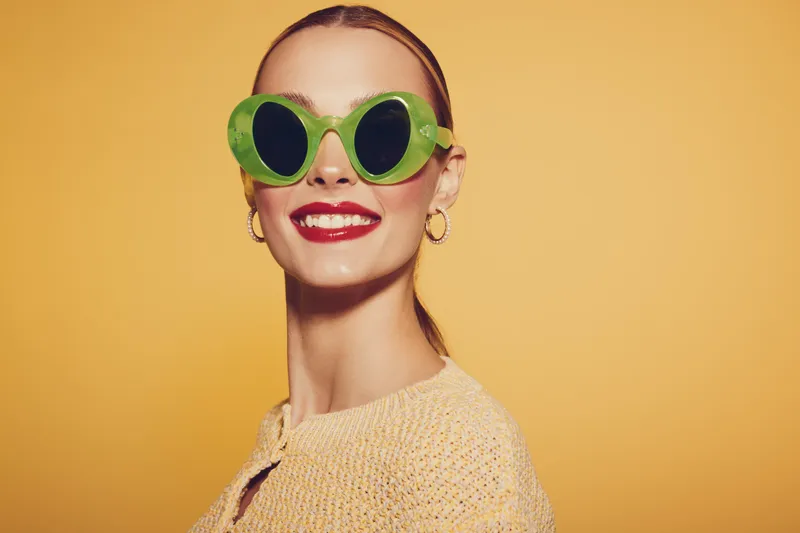 Smiling woman with green sunglasses and red lipstick poses against yellow background. Fashion portrait with bright colors and confident expression. Concept of style and summer mood.