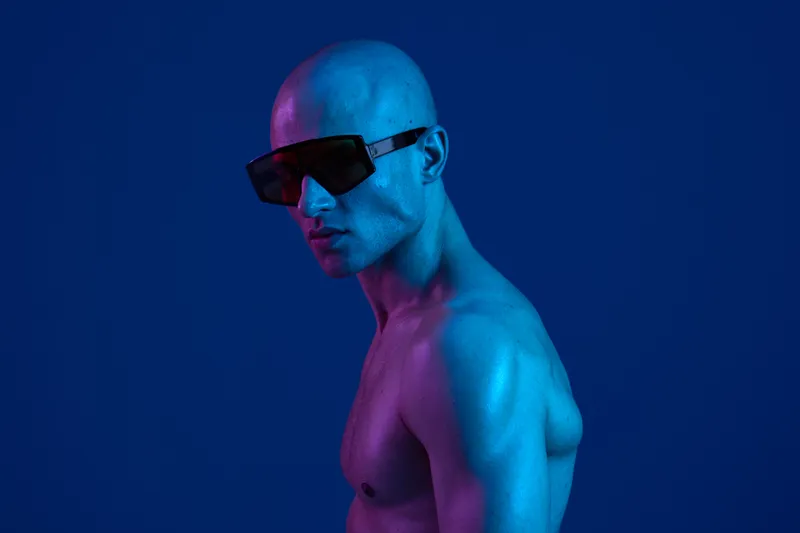 A stylish man with a bald head, wearing bold sunglasses, showcases a vibrant blue lighting effect. The striking contrast highlights his confident expression and unique look.
