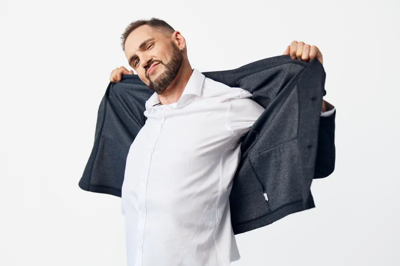 A confident business professional man poses on a clean isolated white background, wearing a white shirt and dark blazer draped over his shoulders, conveying relaxed charm and positive emotion.