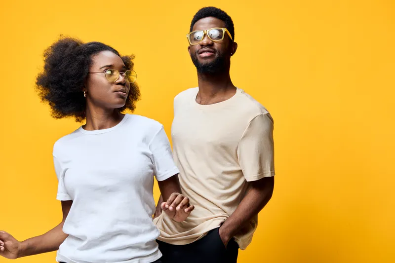 Young African couple wearing sunglasses, smiling against a vibrant yellow background, showcasing youthful energy and fashionable summer style