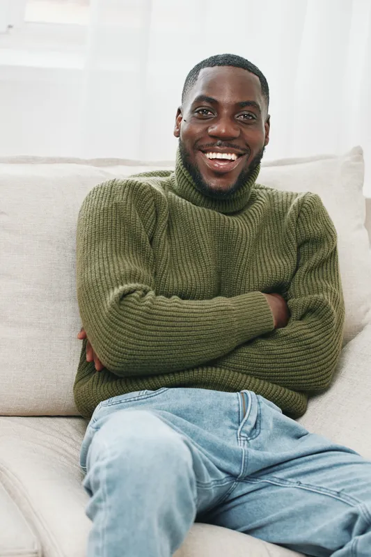 Smiling African American man in a cozy green sweater, sitting on a light sofa, conveying warmth and relaxation, perfect for lifestyle and home themes