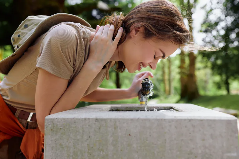 Woman drinking from a fountain with backpack in a park, smiling as she leans over a concrete basin to get water, outdoor summer scene, casual clothes, hydration and nature travel.