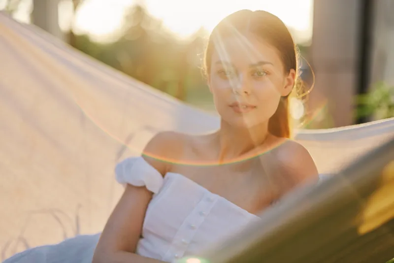 Young woman relaxing in a hammock during sunset, wearing a light summer dress, radiating calmness and serenity in a warm, golden light, ideal for wellness and summer themes