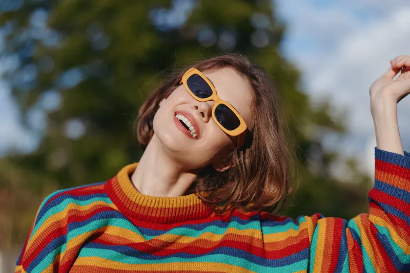 Woman in yellow sunglasses smiling in colorful rainbow sweater, portrait outdoors enjoying sunlight and casual fashion, joyful expression and cozy knitwear style in park setting