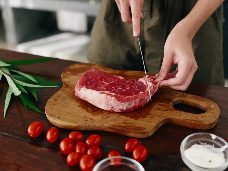 Женщина с ножом в руке нарезает свежий стейк из мяса для запекания на кухне