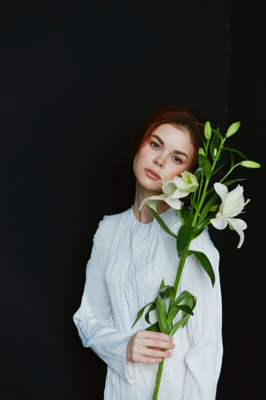 Elegant young woman with lilies, wearing a white dress against a dark background, exuding serenity and grace, showcasing natural beauty and emotion