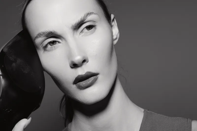 Close-up portrait of a fair skinned Caucasian woman with slicked back hair holding a black shoe near her face, studio lighting.