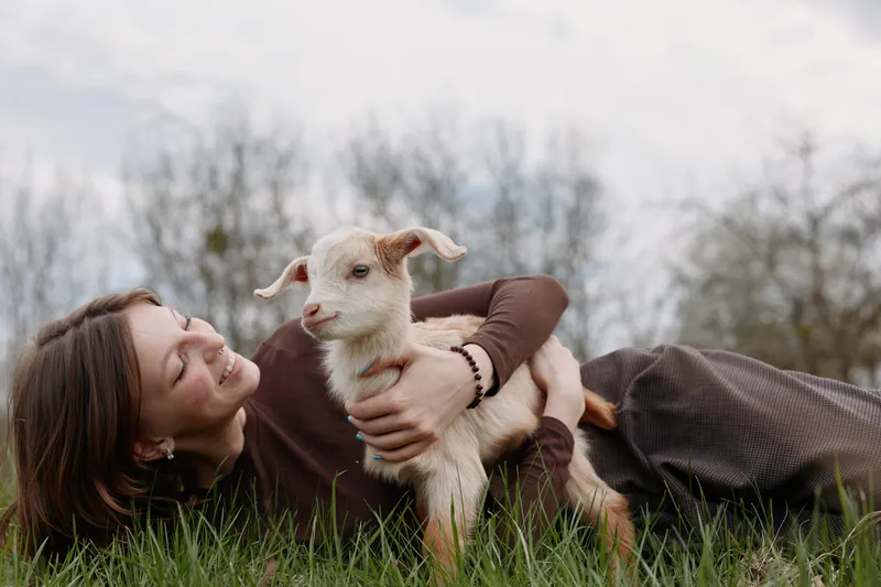 A woman lies on grass outdoors, embracing a young goat kid with a gentle smile, savoring a calm, affectionate moment. A lifestyle scene of companionship and simple rural joy.