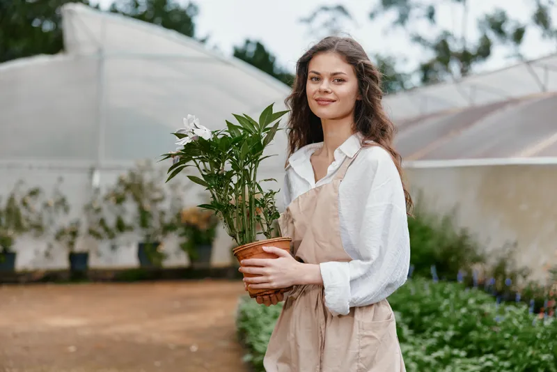 Young woman in light clothing holding a potted plant in a greenhouse, showcasing beauty and serenity while promoting eco friendly living