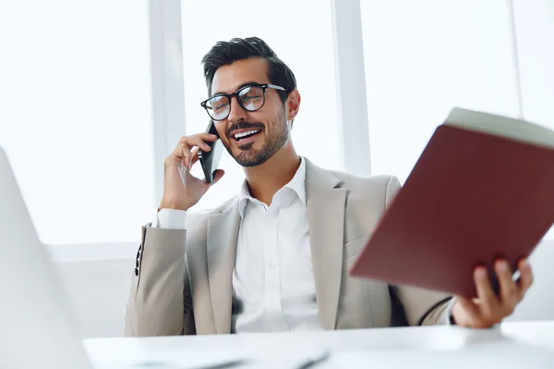 Мужчина в деловом костюме сидит на рабочем месте с ноутбуком и разговаривает по мобильному телефону, держа в руках документы