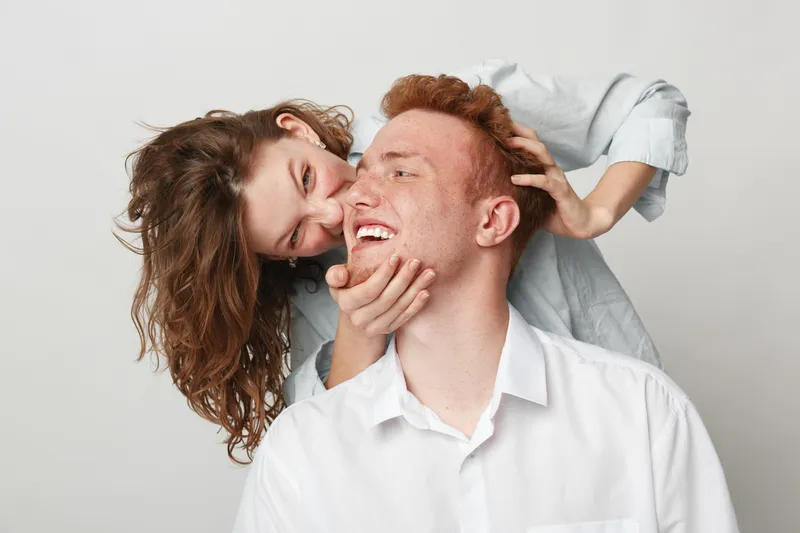 Happy young couple with red hair laughing and playing together on white background. Woman embraces man from behind showing affection and joy. Concept of love and relationship.