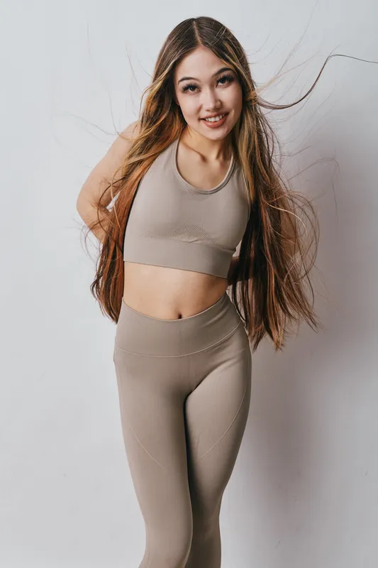 Young woman in seamless beige activewear smiling with long flowing hair, posing with hands behind back, isolated on white background for fitness and lifestyle concept.