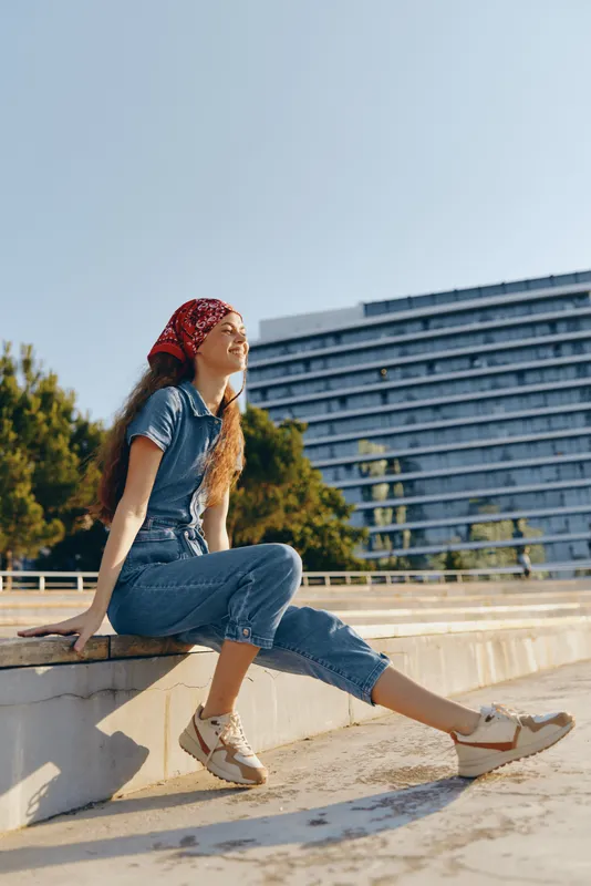 Lifestyle woman in modern boho-western denim street style sits relaxed outdoors with a red bandana, enjoying sunshine and urban scenery with film color tones.