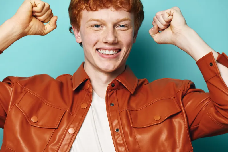 Young man with red hair wearing brown jacket smiling and flexing arms showing strength and confidence against blue background. Portrait of happy male with positive energy.
