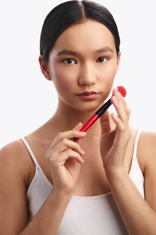 Asian woman applies makeup with a soft brush on the lips in a close up shot. The scene conveys focus, calm concentration, and a radiant, everyday beauty routine.