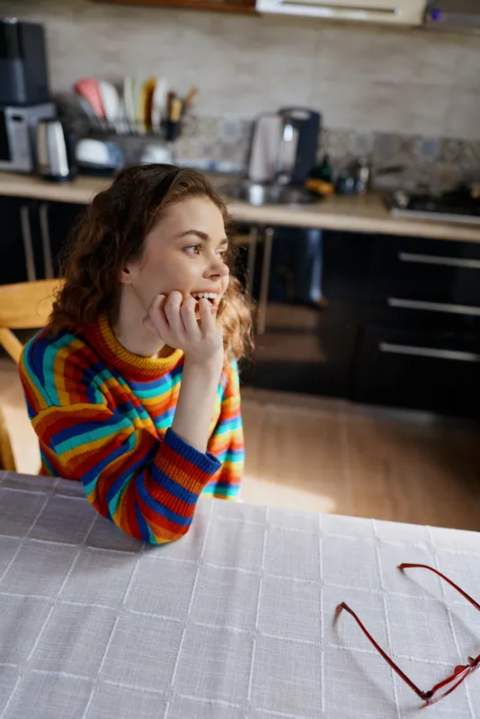 Smiling woman with curly hair in a colorful striped sweater in a cozy kitchen, a warm homey setting. Clear lifestyle concept with everyday routine and soft interior lighting.