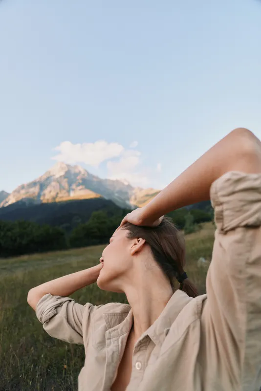 Young woman in a beige shirt stands in a grassy meadow with distant mountains, hands in her hair, exhaling gently as she enjoys a carefree outdoor moment