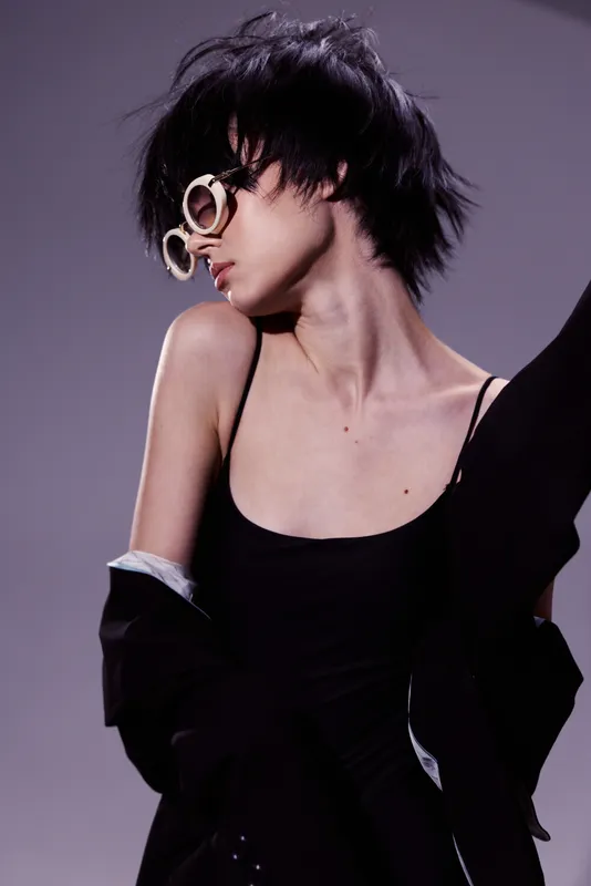 Stylish woman with short dark hair wearing round sunglasses and black outfit in a minimalistic studio setting.