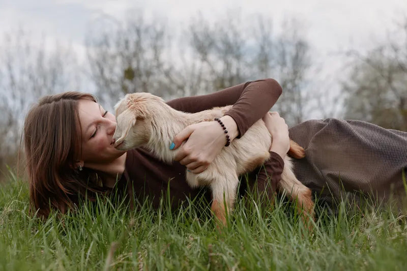 Lifestyle moment of a woman lying on grass outdoors, kissing a young goat kid, tender interaction, rustic natural setting, simple joy and companionship in open countryside field