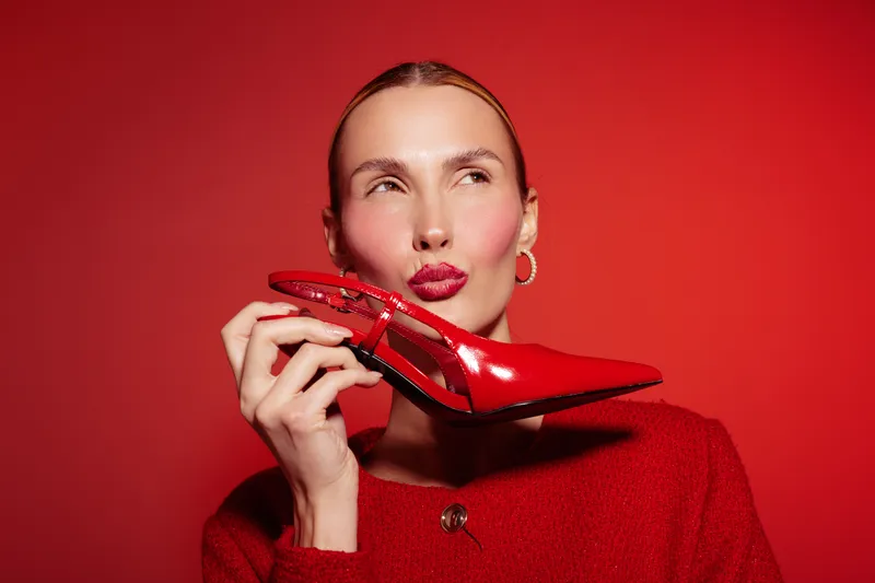 Stylish woman in red sweater holds shiny red high heel shoe near face on red background. Concept of fashion, glamour, and bold style with matching colors and accessories.