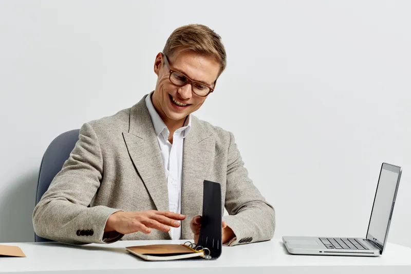 Professional businessman holding a portable speaker, sitting at a desk with a laptop on a plain white background. Office workplace, technology device, communication, modern workspace, casual clothing,