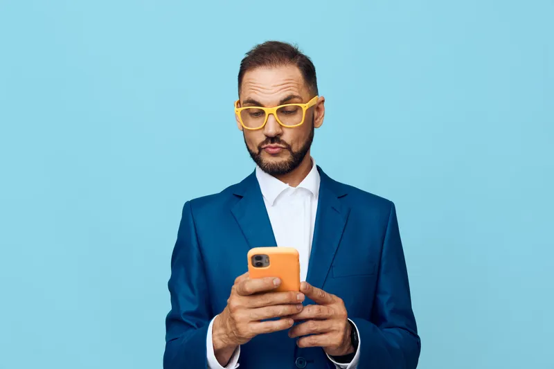 Professional man in a sharp blue suit uses a smartphone, displaying focused emotions of concentration and confidence against a bright isolated colored background.