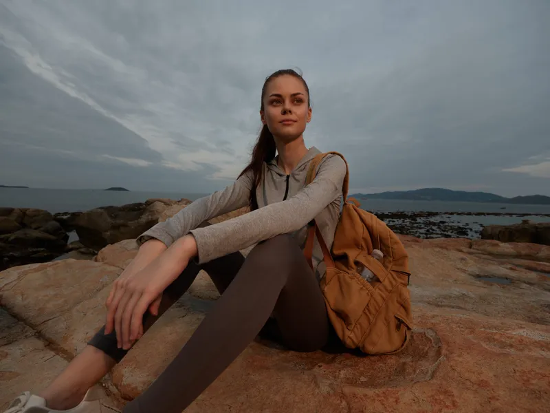 Young woman in activewear with a backpack enjoying nature at sunset on rocky beach, representing outdoor lifestyle and adventure