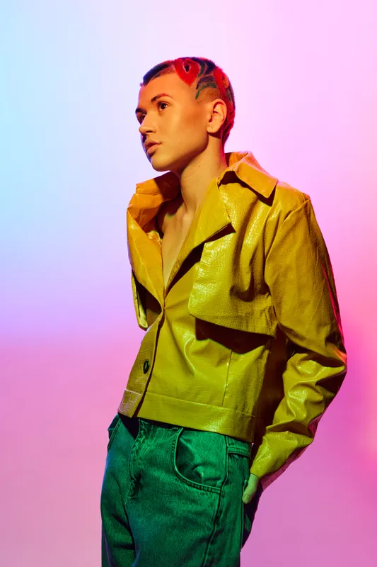 Young woman with artistic short hair wearing bright yellow jacket and teal pants, standing against vibrant pink and blue gradient background in fashionable pose.