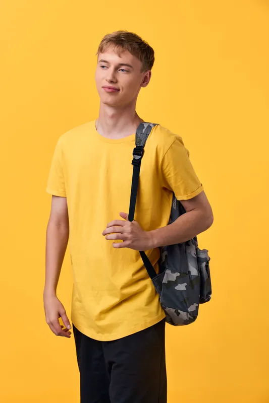 Young man wearing a bright yellow t shirt with a camouflage backpack against a vivid yellow backdrop, casual style, ready for class or travel, relaxed pose, everyday fashion and youth energy.